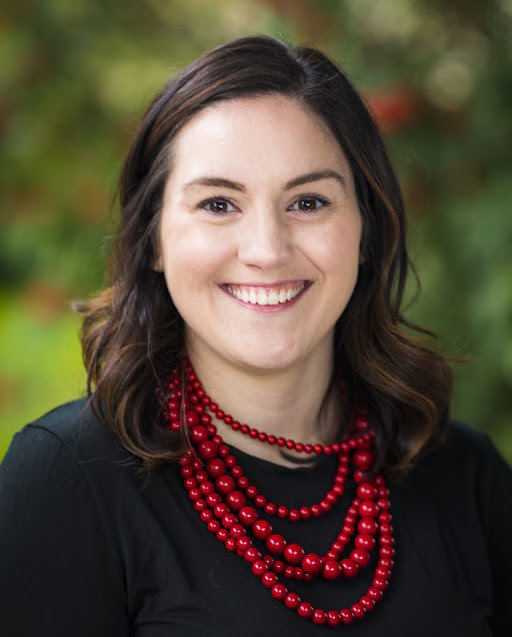 a woman with straight brown hair and a chunky red necklace smiles
