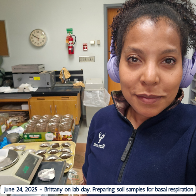 Brittany Kuzma in a lab, wearing headphones and preparing soil samples for basal respiration testing, with labeled jars and equipment behind her.