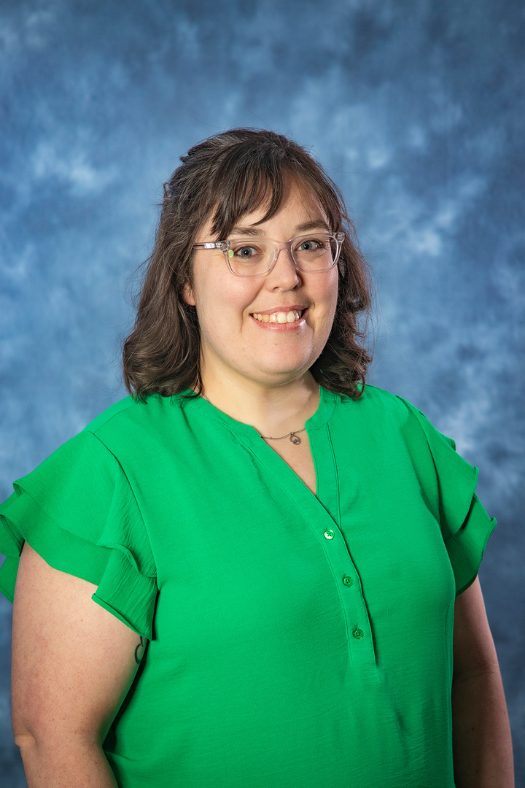 A woman with brown hair and a green top smiles in front of a blue portrait background