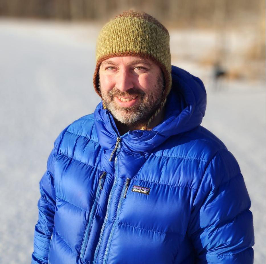 A man with brown hair and beard is wearing a green, knitted, wool hat and blue down jacket