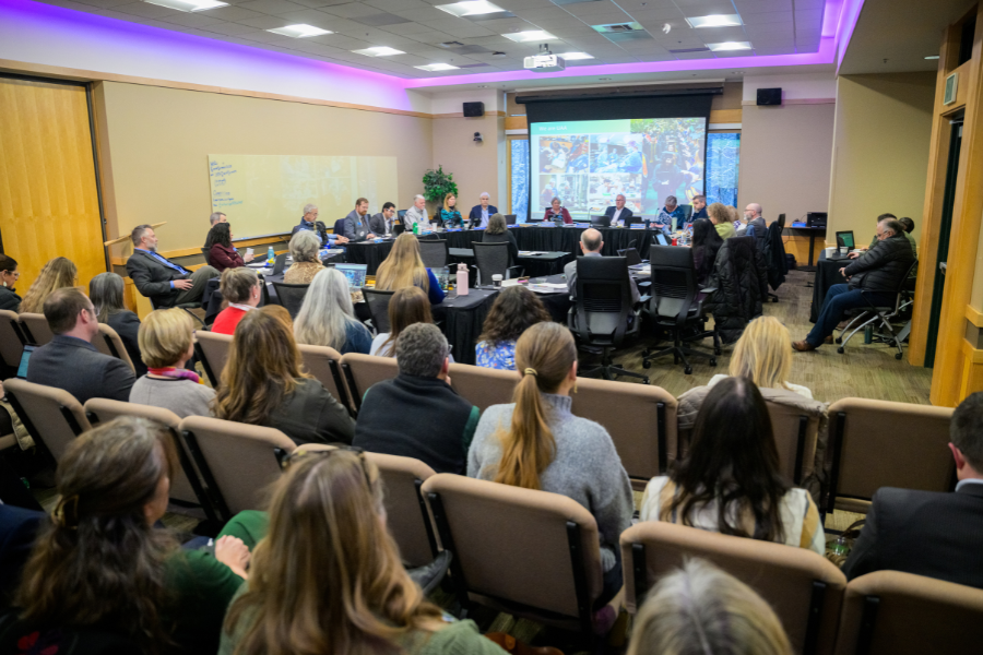 Audience members and University of Alaska Board of Regents gather in a conference room during a meeting, with regents seated at tables facing a presentation screen at the front of the room.