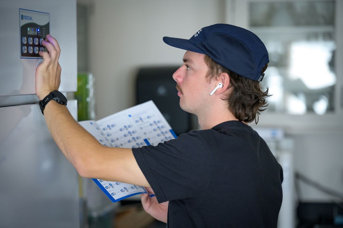 young man in blue hat reaches above to press a button on large machine