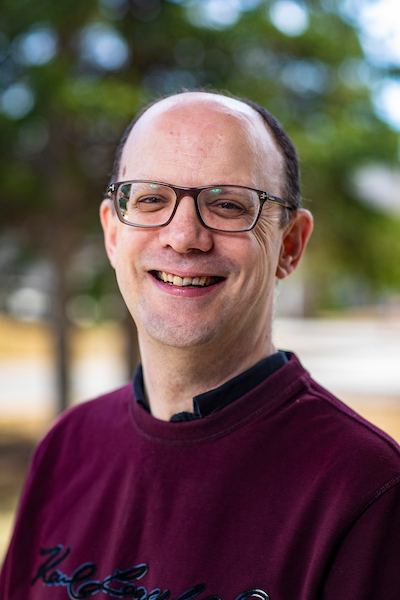 A man with black glasses and maroon shirt smiles in front of a forest background.