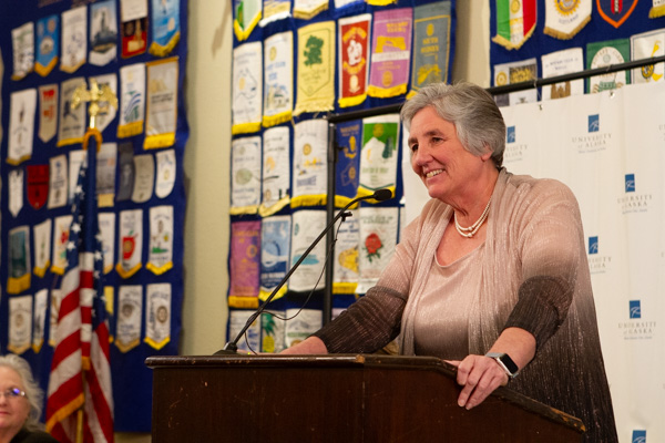 a woman with a pink top and grey hair stands at a podium giving a speech into a black microphone