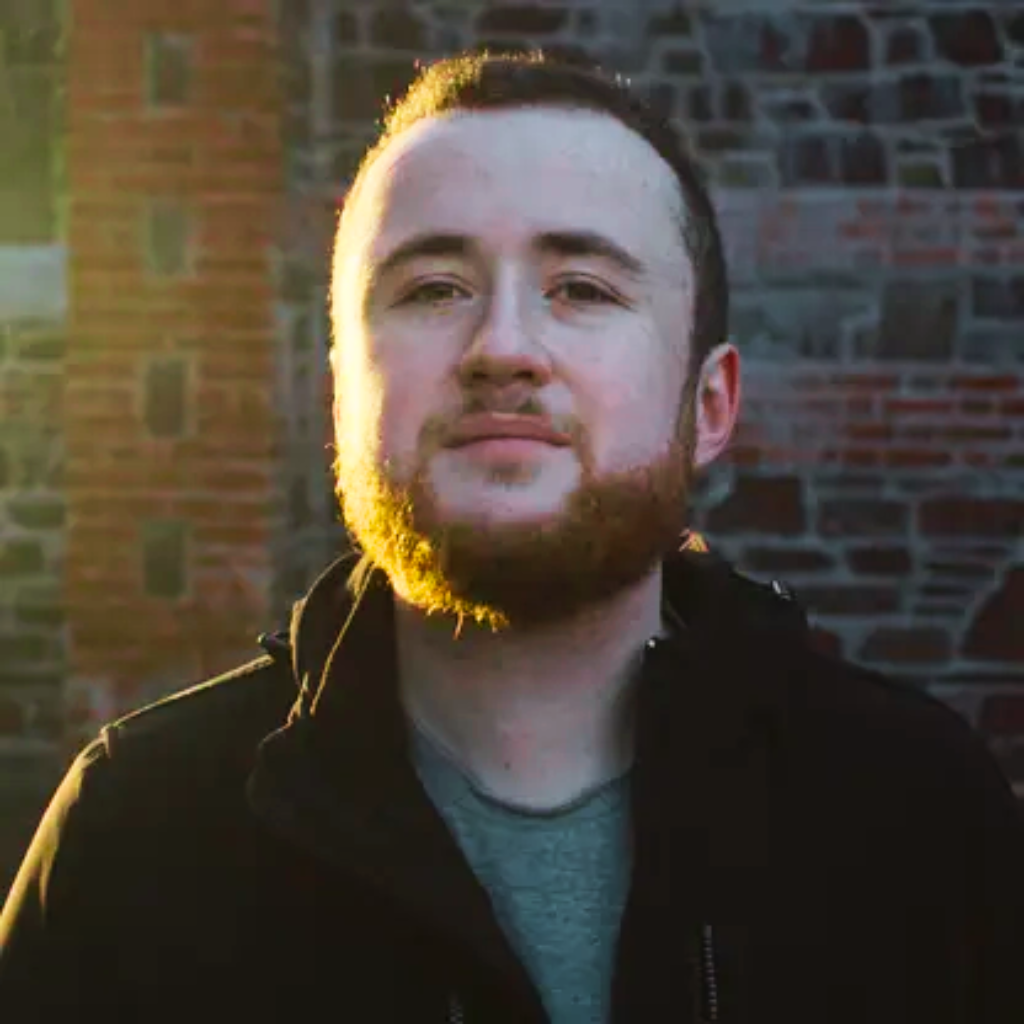 Helperbird founder, Robert James Gabriel, a young white male with ginger hair, beard, and mustache, smiling thoughtfully while gazing at the camera. He is dressed in casual attire and stands in front of an old brick structure in the soft morning light