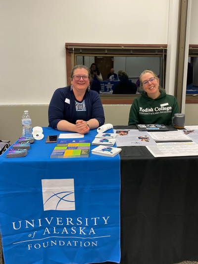 two women sit behind a booth, with blue table cover that reads "UA Foundation"
