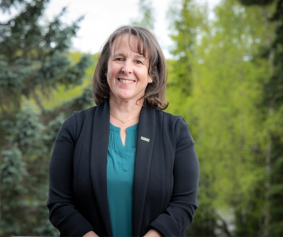 a woman with long brown hair poses in front of an evergreen background