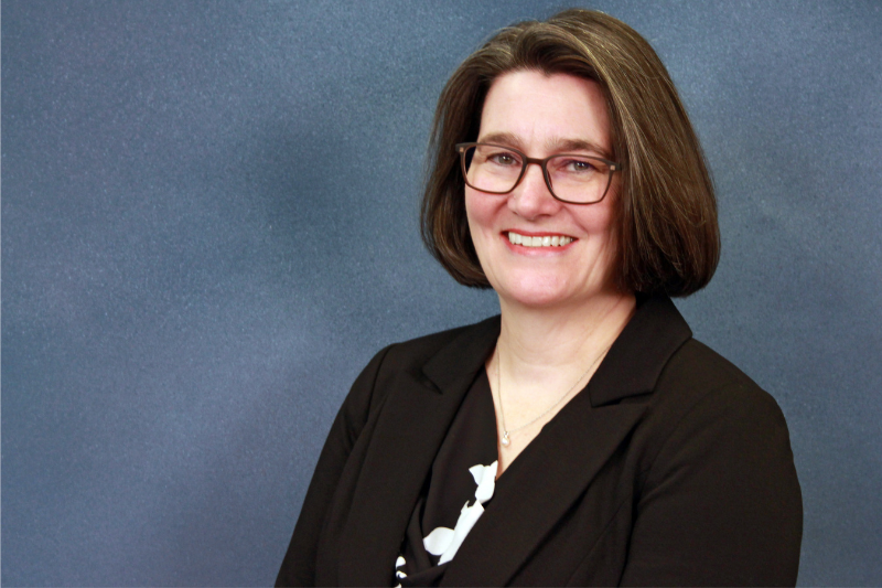 A headshot of a smiling woman with medium-length brown hair and glasses, wearing a black suit jacket.
