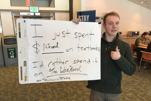 A young male student holds a large white sign and gives a thumbs up