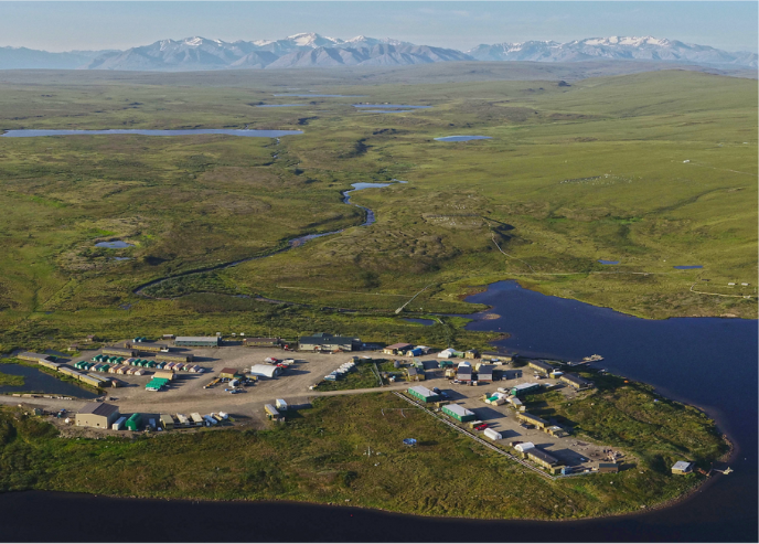 An aerial view of green, summer tundra with a small collection of buildings and a mountain range in the foreground
