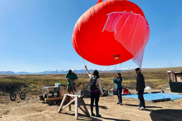 People preparing a large red weather balloon for launch in a field with a mountainous backdrop.
