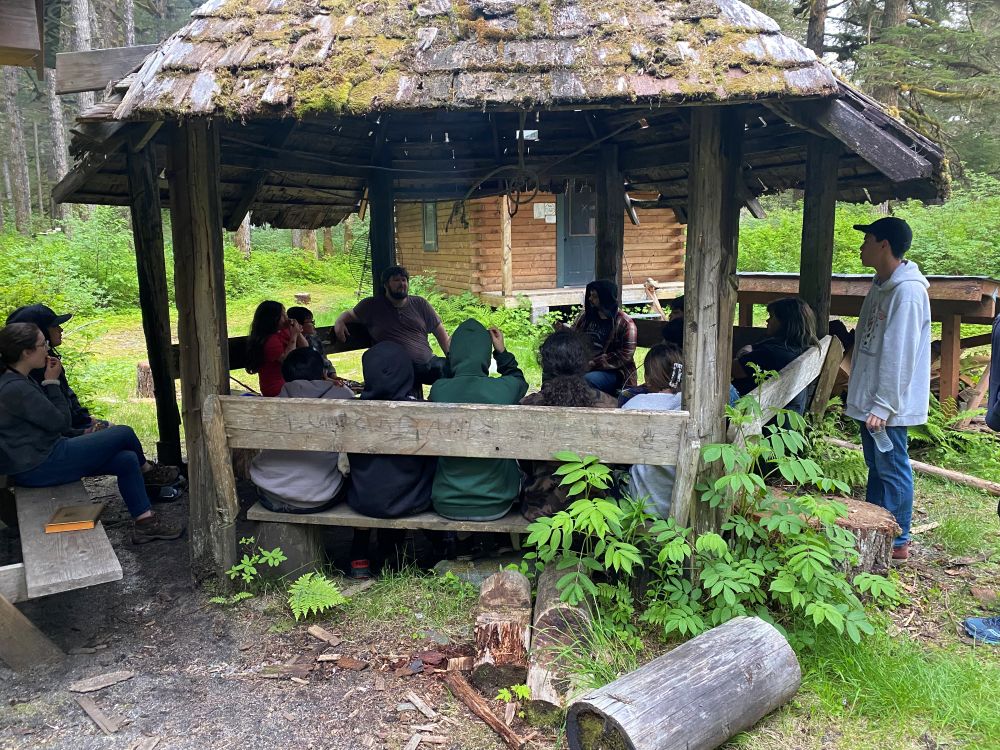 People gather in an outdoor wooden pavilion in a forested area. 