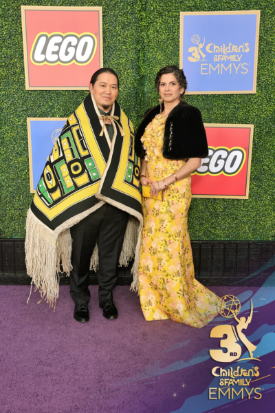 Two individuals posing at the 3rd Children’s & Family Emmys, with LEGO and other sponsors’ logos in the background. One person is wearing a vibrant robe with building-like patterns and the other is in a yellow floral dress with a black shawl.