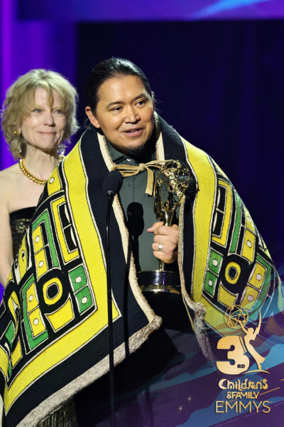 Person holding an award on stage at the Children's & Family Emmys, wearing a traditional indigenous blanket, with another person in the background smiling.