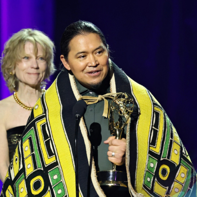 Twitchell, with black hair and a traditional robe stands on stage, holding an Emmy award and speaking into a microphone. An older woman with short brown hair stands behind him.