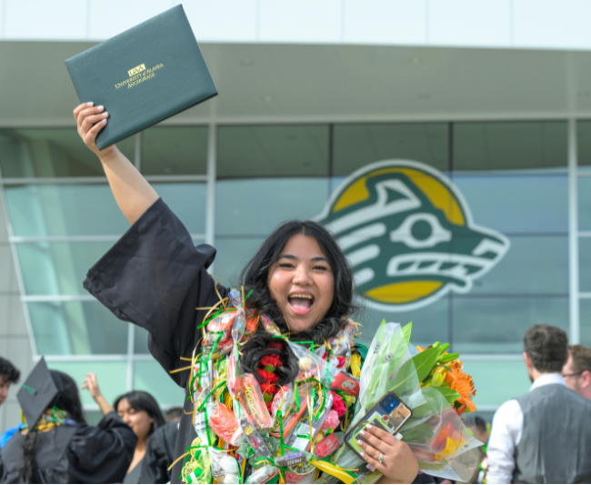 A young girl with dark hair, wearing graduation regalia holds up her UAA diploma in front of a UAA building with a crowd behind her