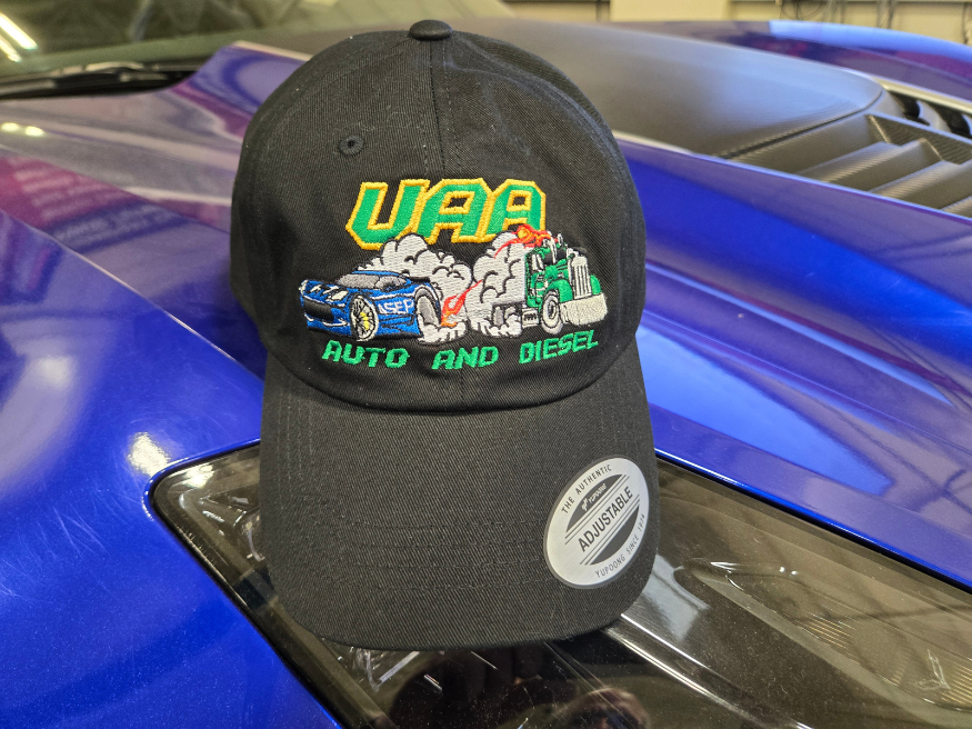 A black UAA Auto and Diesel embroidered cap rests on the hood of a blue vehicle, featuring a custom design of a sports car and semi truck.