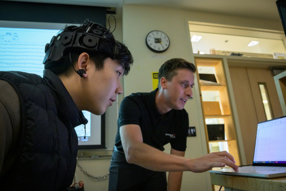 A student wearing a black EEG headset with an ear clip leans toward a laptop as a trainer points to the screen, with projected brain-wave readings in the background.