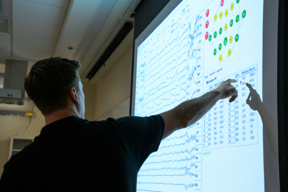 A person points at a projected screen of EEG software showing rows of wavy brain-activity readings, a color-coded map of sensor positions on the head and a table of signal values.