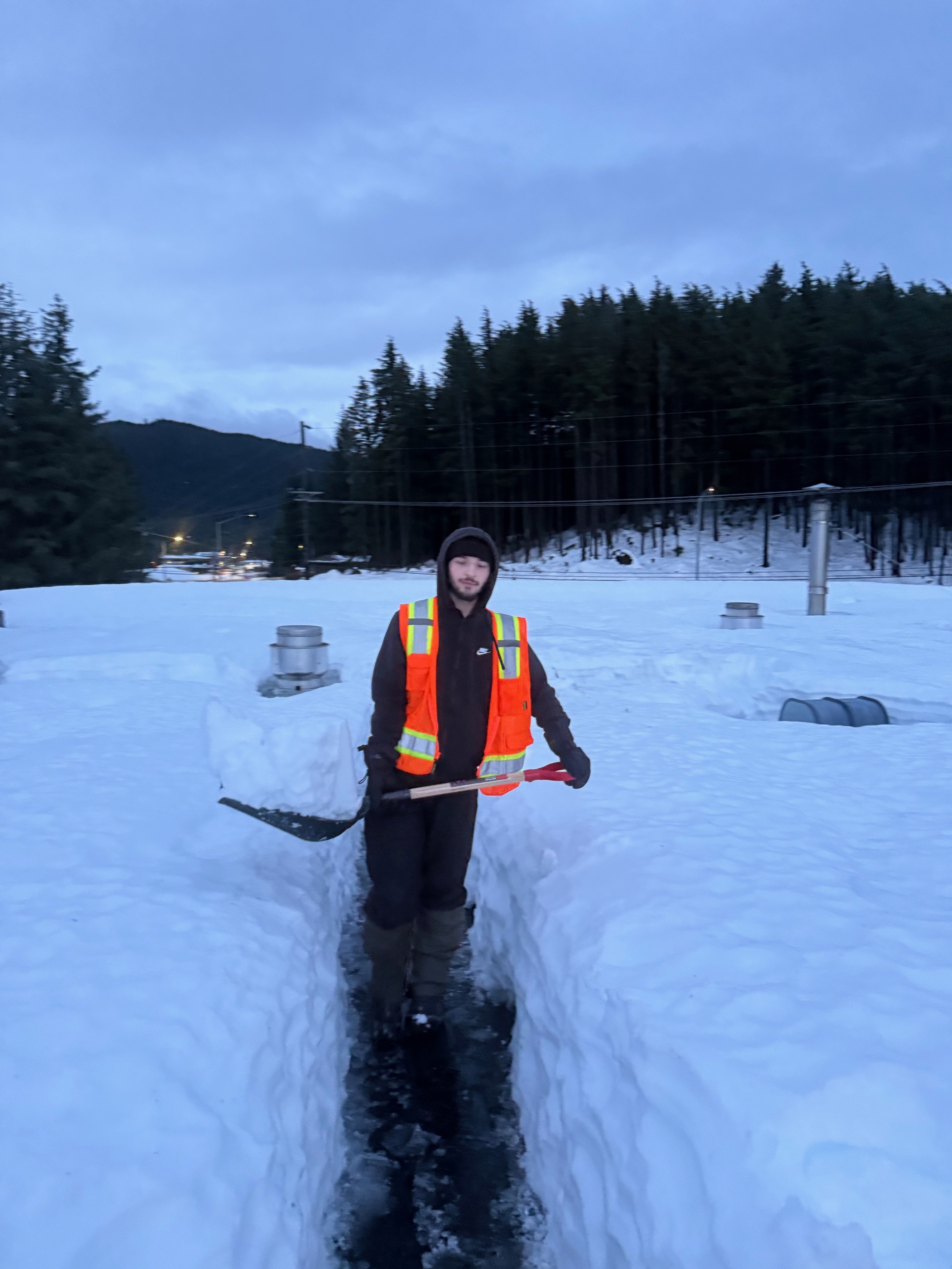 A person in an orange fest walks through a tunnel in waist-deep snow, carrying a shovel full of snow.