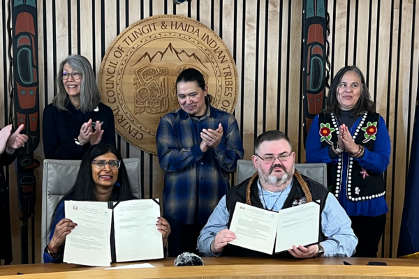 Two people hold up signed documents while six people applaud behind and beside them. They are flanked by the Tlingit & Haida seal, two formline posts, and the American, Alaskan, and Tlingit & Haida flags.