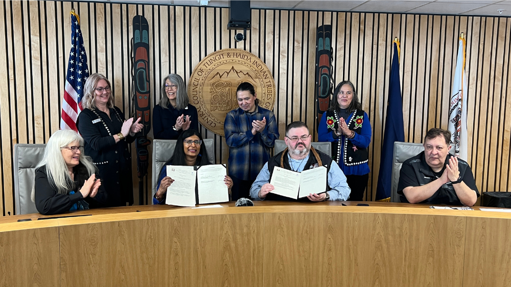 Two people hold up signed documents while six people applaud behind and beside them. They are flanked by the Tlingit & Haida seal, two formline posts, and the American, Alaskan, and Tlingit & Haida flags.