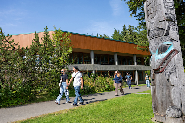 A group of students walk by a totem pole on a sunny day on campus