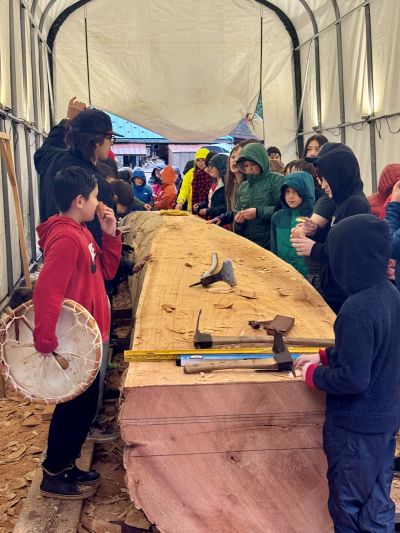 Children stand around a large log with hand tools and wood chips resting on it.