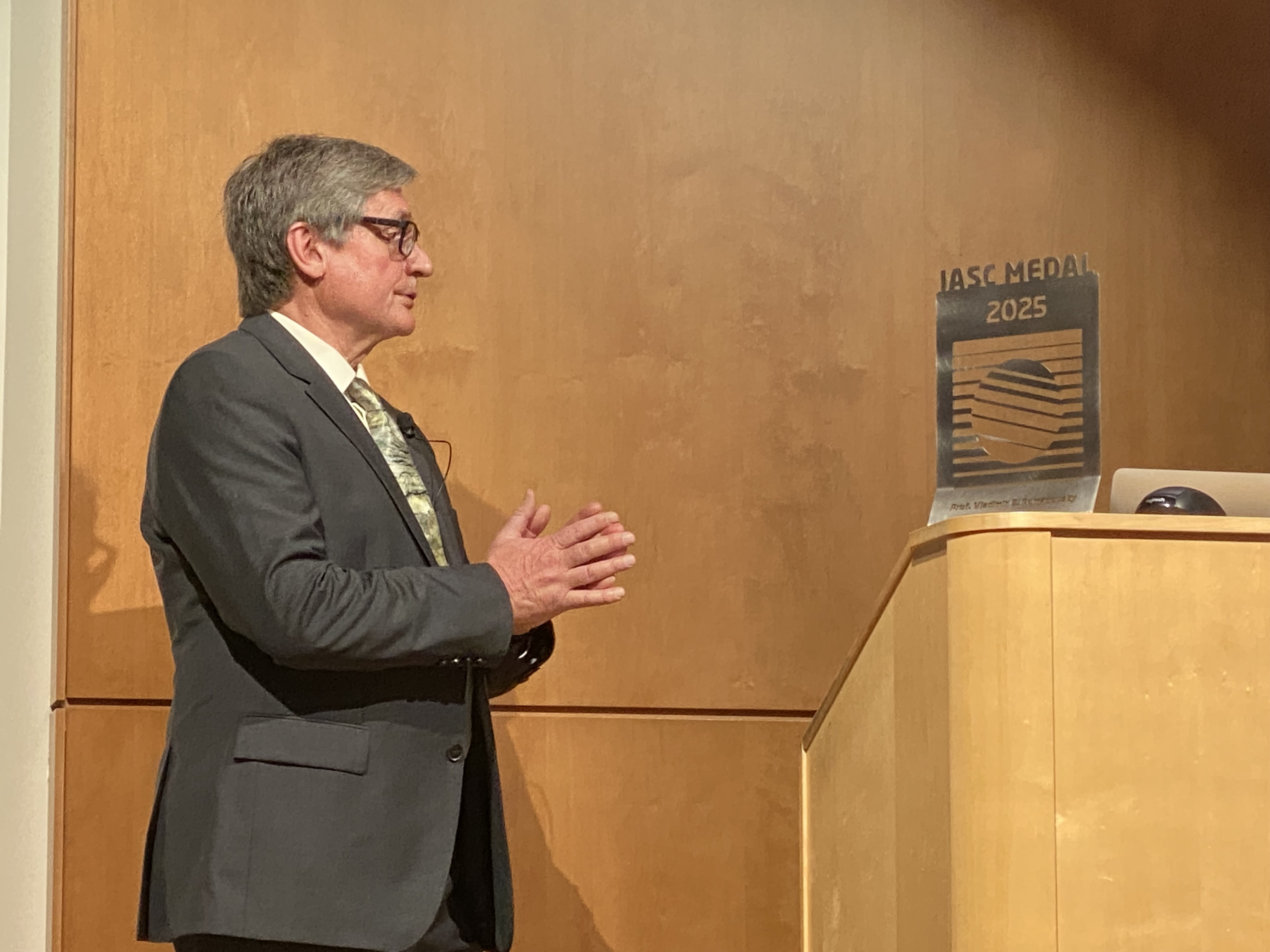 A man with grey hair and grey suit stands next to a podium with his hands clasped. On the podium is a crystal and wood award.