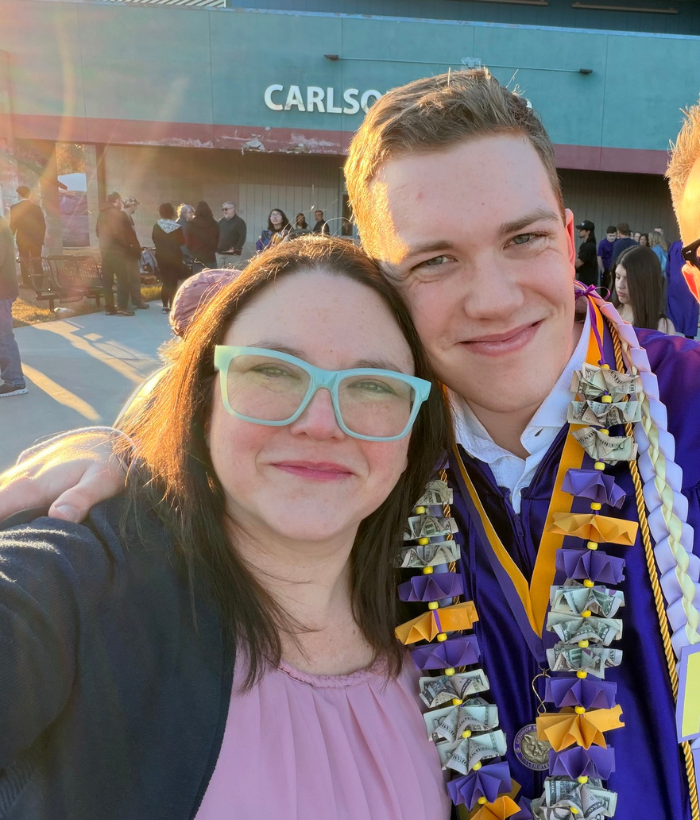 Two people pose for a selfie outside a building during an outdoor graduation event. One person is wearing a purple graduation gown adorned with leis made of folded dollar bills and purple and gold ribbons. Groups of people are gathered in the background near the building entrance in warm evening sunlight.