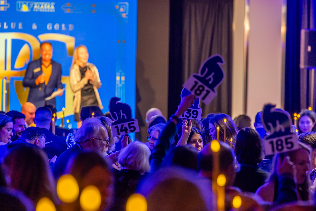 Gala guests raise UAF polar bear-shaped bidding paddles during a live auction, with two emcees visible on a blue and gold illuminated stage in the background.