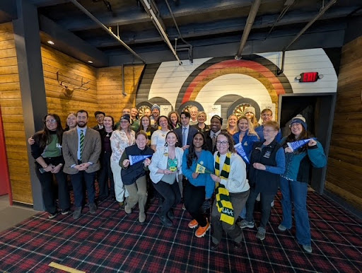 a large group of people pose in a room with wooden walls and plaid carpet