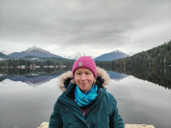 A woman with blonde hair wears a pink hat and jacket in front of a body of water with mountains in the background.