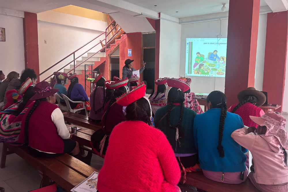 Participants in Peru sit on wooden benches during a community health education session while a presenter points to a projected slide at the front of the room.