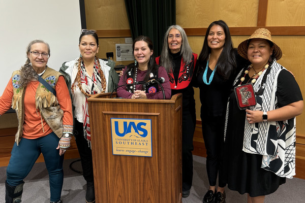 Six people stand behind a University of Alaska Southeast podium during an event. Several wear traditional Alaska Native regalia or accessories, including woven hats, beaded jewelry and patterned garments. The group poses together in a meeting room with wood-paneled walls and a projection screen in the background.