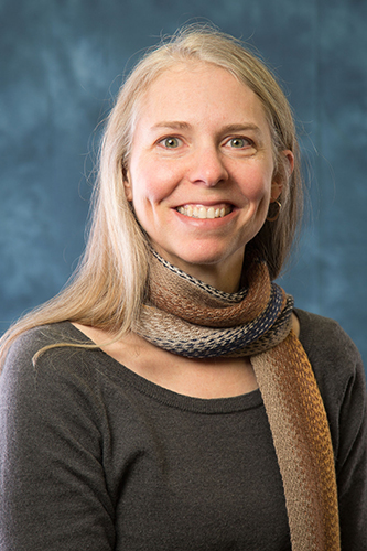 A woman with long, grey hair and black shirt smiles in front of a blue portrait backdrop.