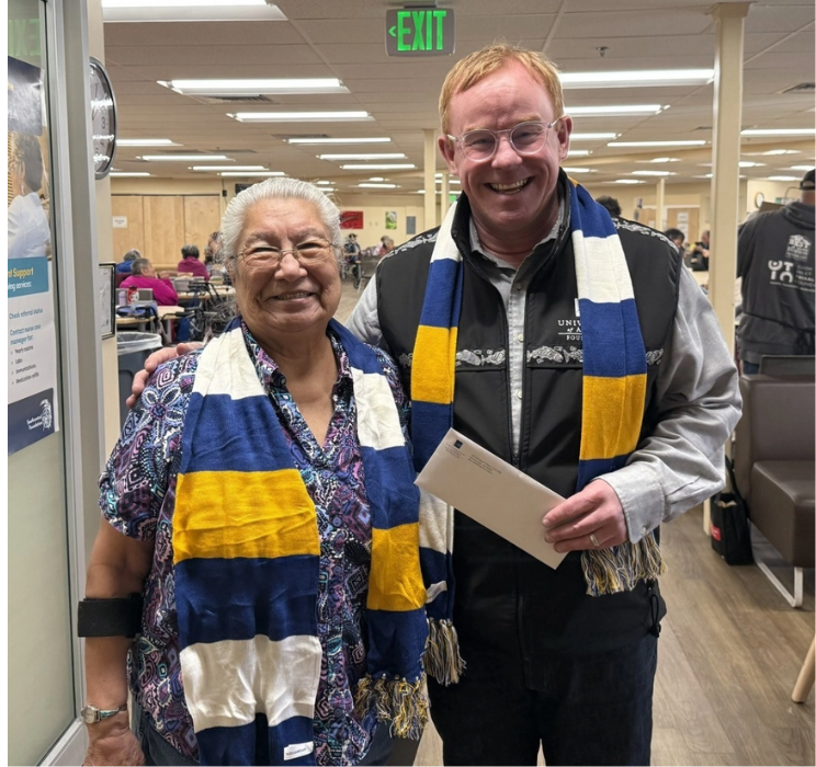 Myrtle Vandoorn and Harry Need standing together in the Southcentral Foundation Elders Program wearing blue and yellow UAF scarves.