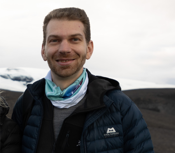 Portrait of Magnus de Witt, a man with short brown hair wearing a dark puffer jacket and teal neck gaiter, smiling outdoors with a snow-covered landscape visible behind him.  