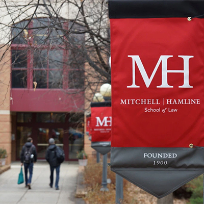students walk on sidwalk in front of a stone building and the red lightpost sign with Mithcell Hamline School of Law hangs close to the camera