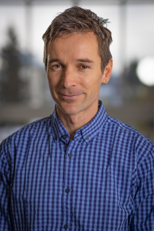 Headshot of Pascal Buri, a man with short dark hair wearing a blue gingham button-down shirt, smiling slightly against a softly blurred indoor background.