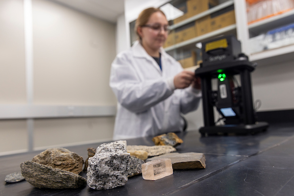 A researcher in safety glasses and a lab coat uses a handheld analyzer to test rock samples in the UAF critical minerals lab