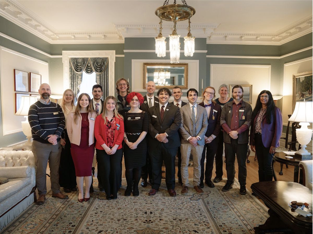 A group of approximately 14 people pose for a photo in a formal room with ornate crown molding, a chandelier, and classic furnishings.