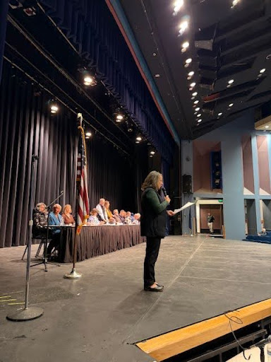 UAA Chancellor Cheryl Siemers addresses the crowd on stage holding a microphone while a panel sit at a long table behind her
