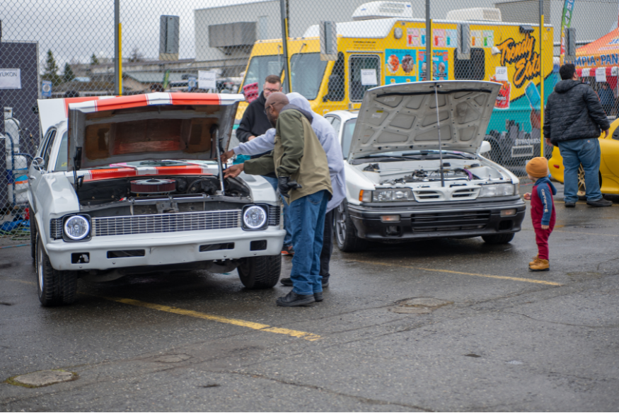 Attendees examine vehicles with open hoods at the 2026 UAA Auto Expo in Anchorage, with food trucks visible in the background.