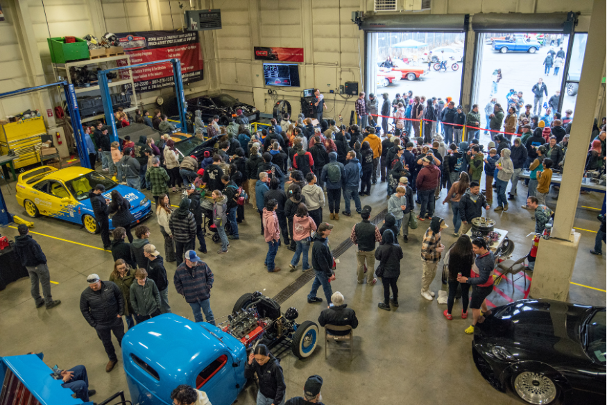A large crowd fills the UAA Automotive and Diesel Technology building during the 2026 Auto Expo in Anchorage, with custom vehicles on display including a blue hot rod in the foreground.