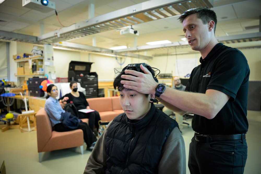 A trainer fits a black EEG headset onto a seated student's head as others watch from a couch in the lab.