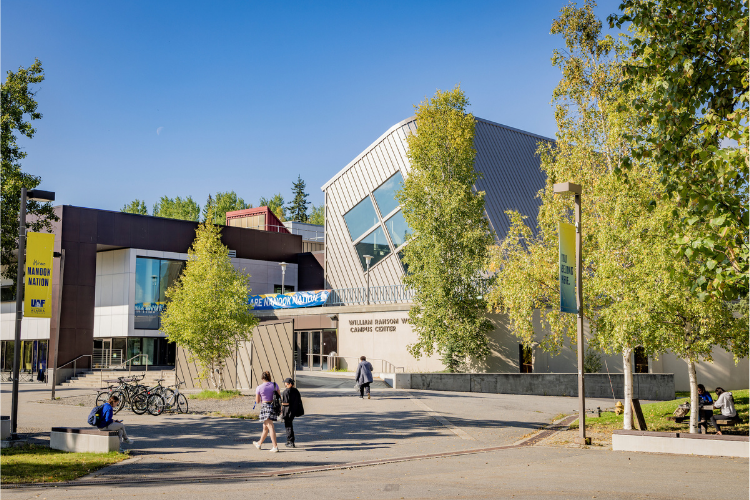 The William Ransom Wood Center on the University of Alaska Fairbanks campus on a sunny day. Trees line a pedestrian walkway leading to the modern building, while several people walk across the plaza and bicycles are parked nearby.