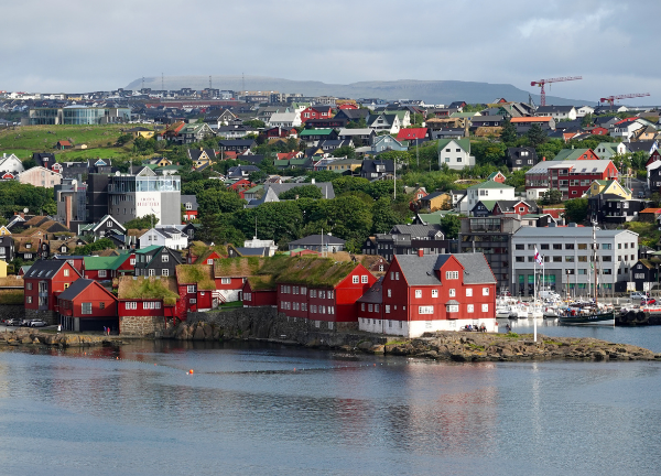 A view of Tórshavn, capital of the Faroe Islands, showing colorful buildings with traditional turf roofs along the harbor waterfront, with residential neighborhoods climbing the hillside behind them.