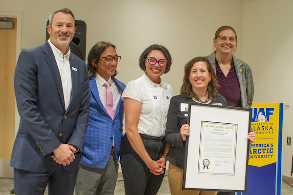University of Alaska Chief Finance Officer Luke Fulp, Alaska Sen. Scott Kawasaki, Alaska Rep. Maxine Dibert, Lael Oldmixon, executive director of Education Trust of Alaska, Alaska 529 and UA Scholars, and Alaska Rep. Ashley Carrick pose with a framed legislative citation during a recognition event. Oldmixon holds the citation while the group stands beside a UAF podium.