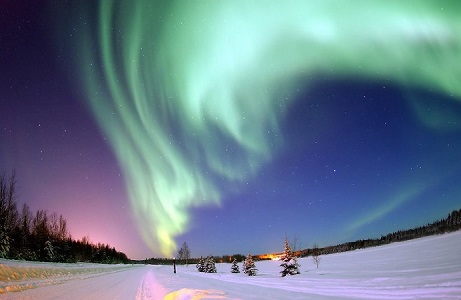 Green aurora borealis in the sky over a snow covered road. The sky takes up much of the image, and it is pink and purple to the left side of the aurora, and dark blue on the right side. There are trees lining the snowy road, and the snow is reflecting orange and pink light.